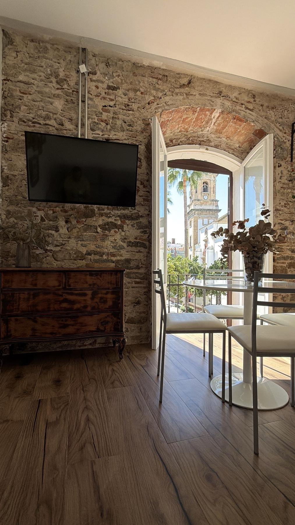 Salón con pared de piedra, cómoda restaurada y mesa redonda junto a un balcón con vistas a una torre